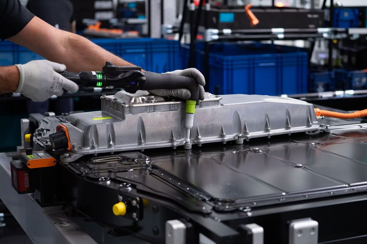 Gloved hands use a specialized tool on a large metallic battery module, likely in a factory setting.