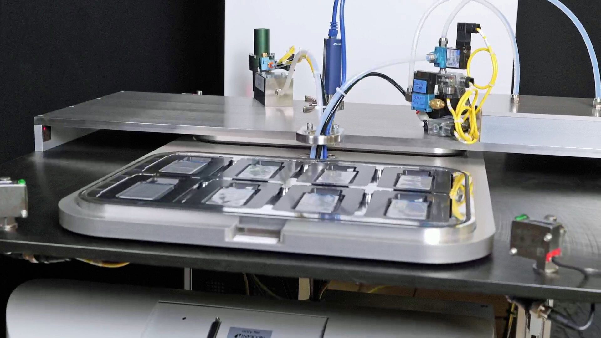 Automated lab equipment featuring a tray of rectangular samples, connected by tubes, wires, and valves.