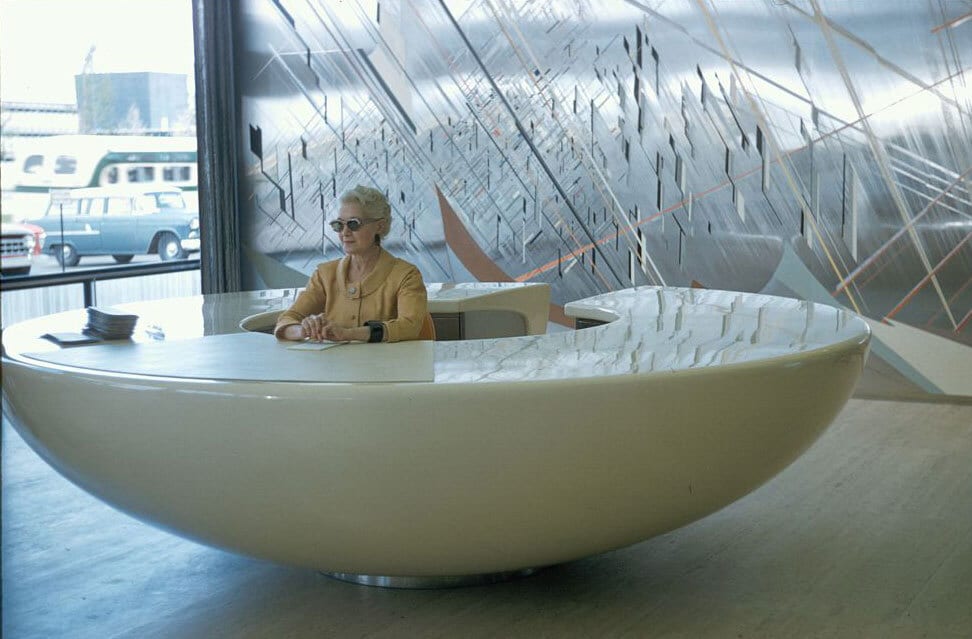 Woman in sunglasses at a curved white reception desk with an abstract art background.