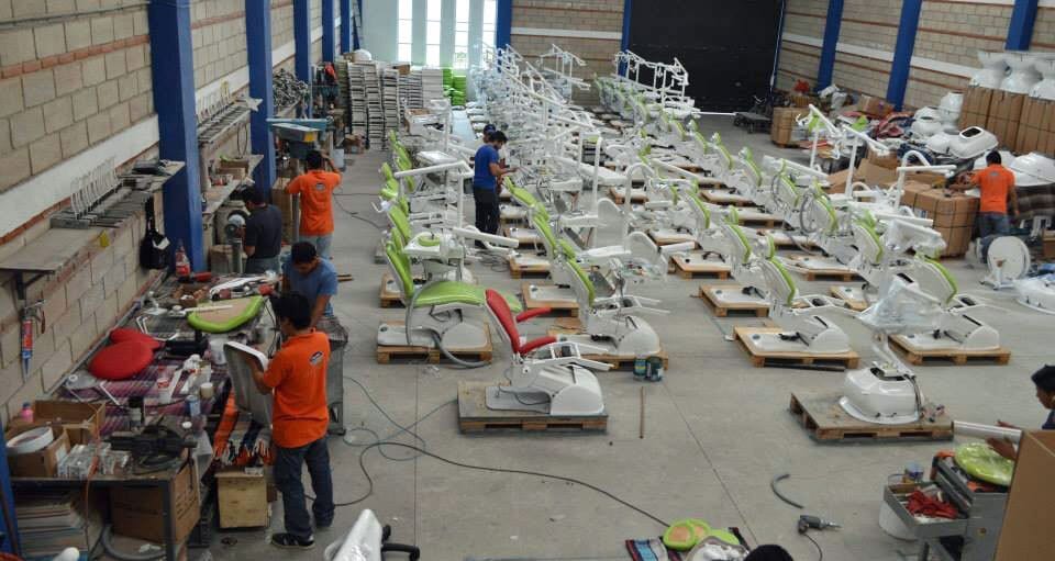 Factory floor where workers assemble dental chairs with green and red upholstery, showing a production line.