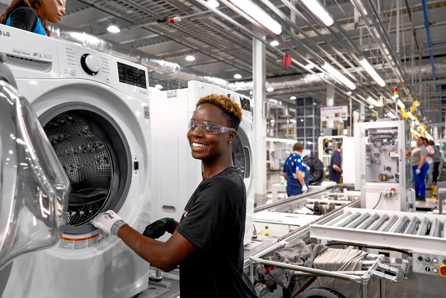 Smiling Black woman in safety glasses assembling an LG washing machine in a factory.