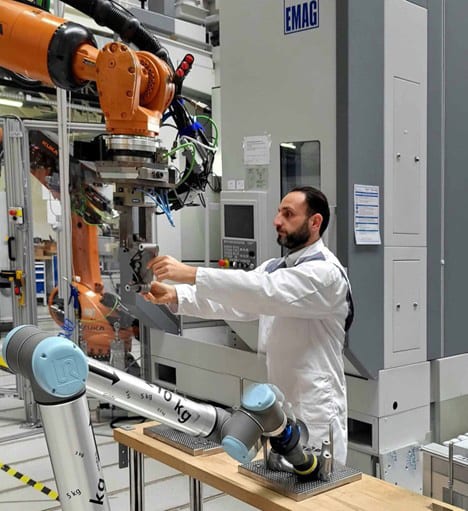 Man in lab coat interacting with an industrial robot, with a collaborative robot and machinery in the background.