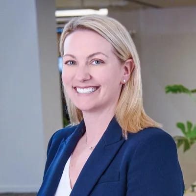 A smiling blonde woman in a navy blue blazer looks confidently at the camera in a professional setting.