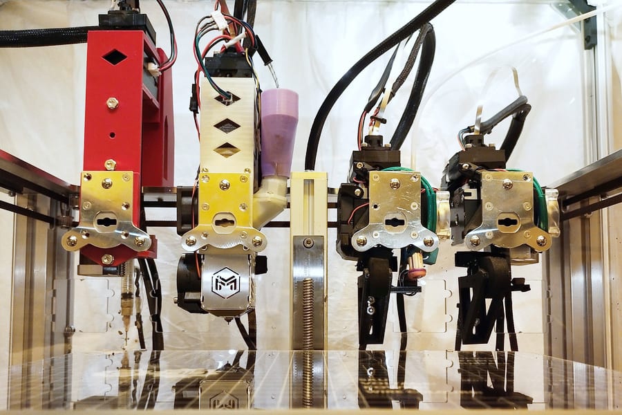 Multiple 3D printer print heads in a row, showing various tool heads and wiring.