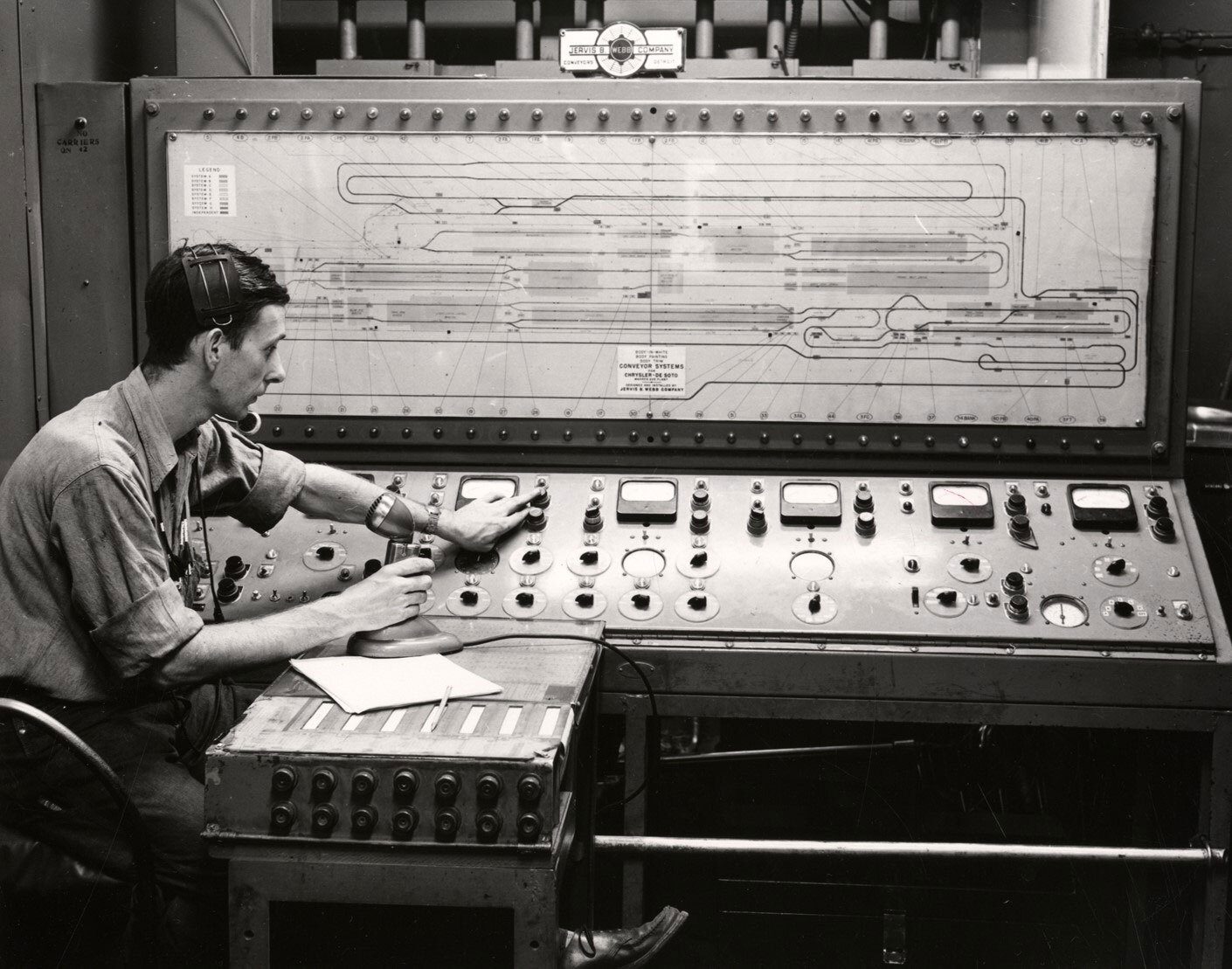 Man in headset operates a vintage control panel displaying a conveyor system diagram.