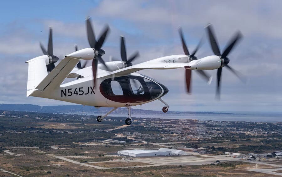 White eVTOL aircraft with blurred propellers flying over a city and bay.