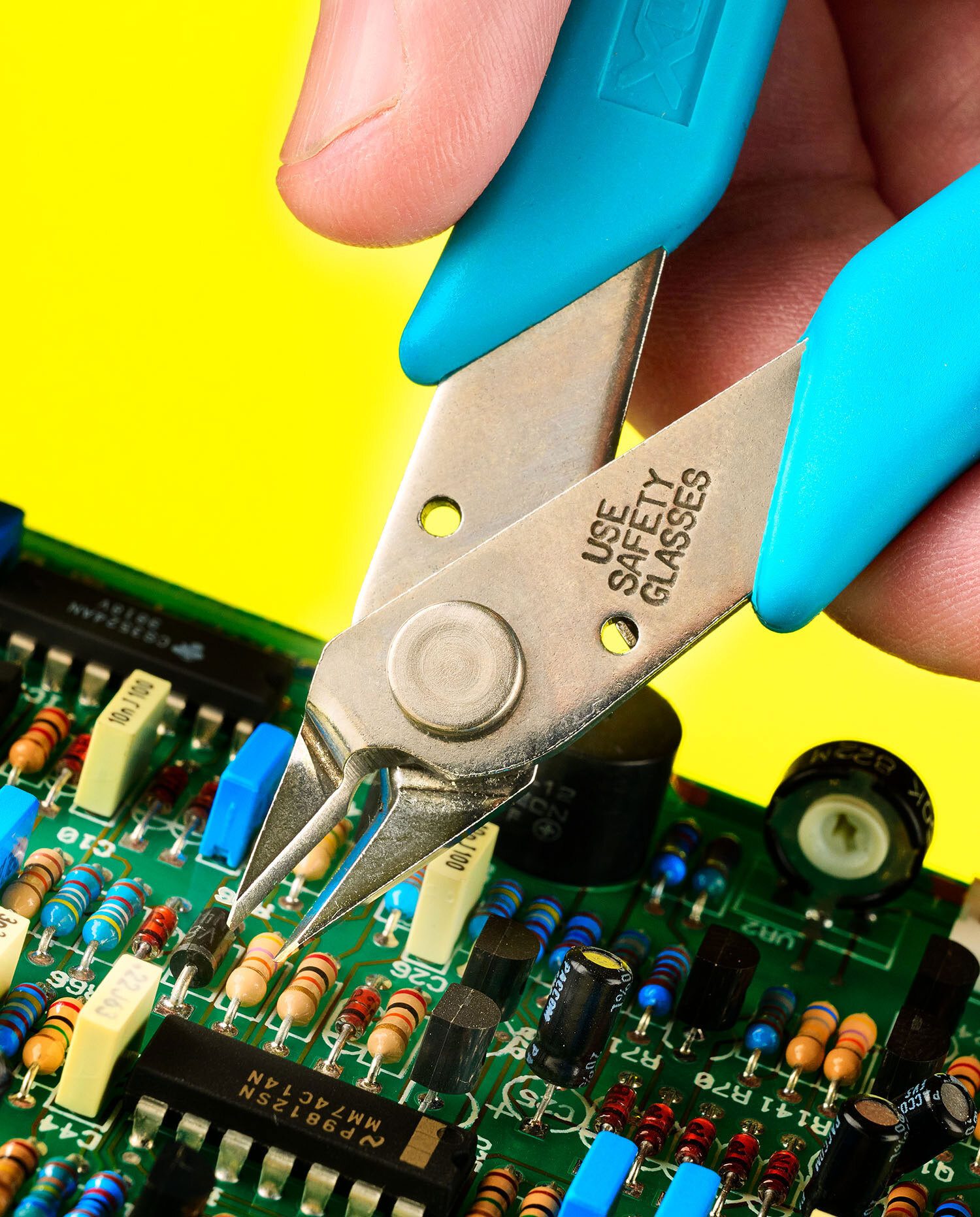 Hand with blue wire cutters ("USE SAFETY GLASSES") positioned to cut a circuit board component.