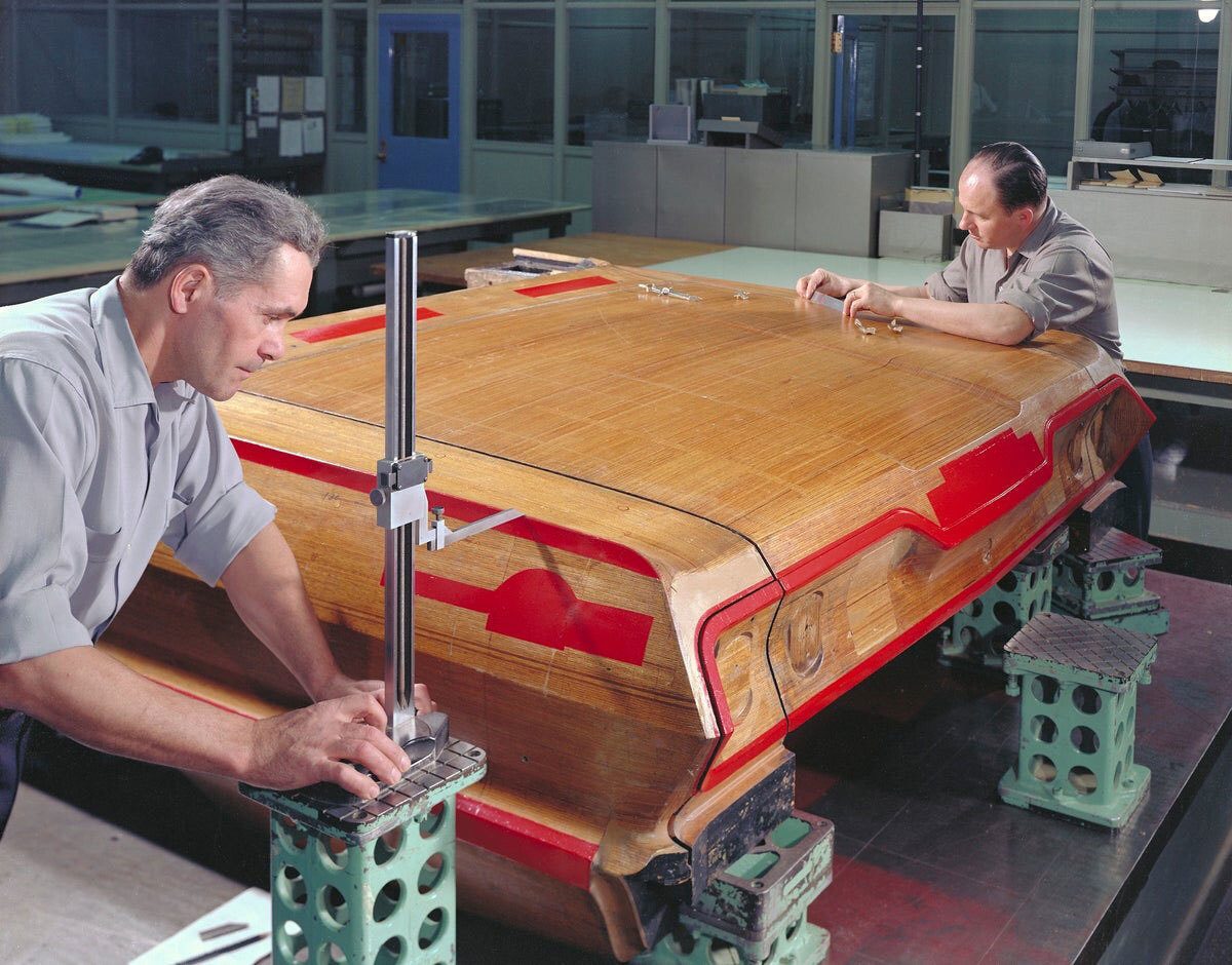 Two men working on a large wooden prototype car model, one measuring with a height gauge, the other inspecting it.