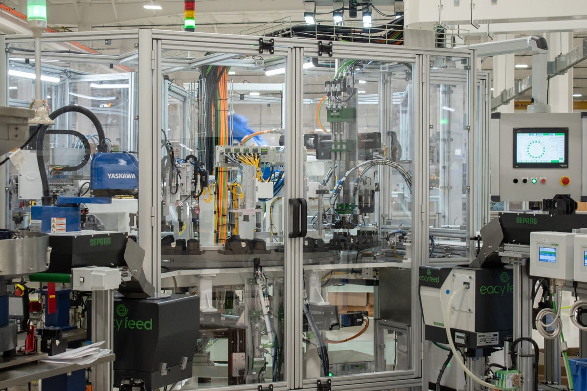 Automated manufacturing line with a Yaskawa robot, control panels, and clear safety enclosure.