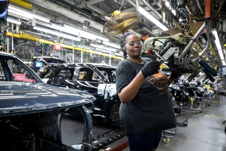 Black woman in safety glasses and gloves works on an assembly line with car bodies.