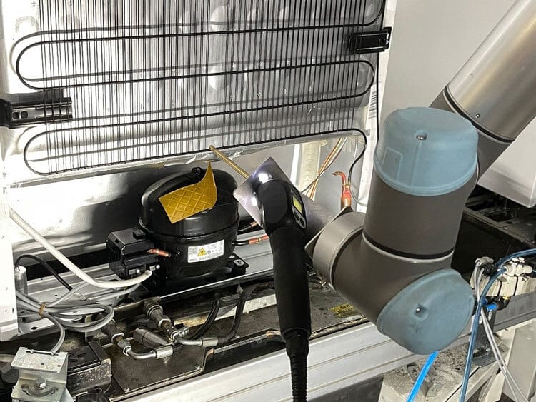 Robotic arm working on refrigerator compressor and condenser coils with a tool.