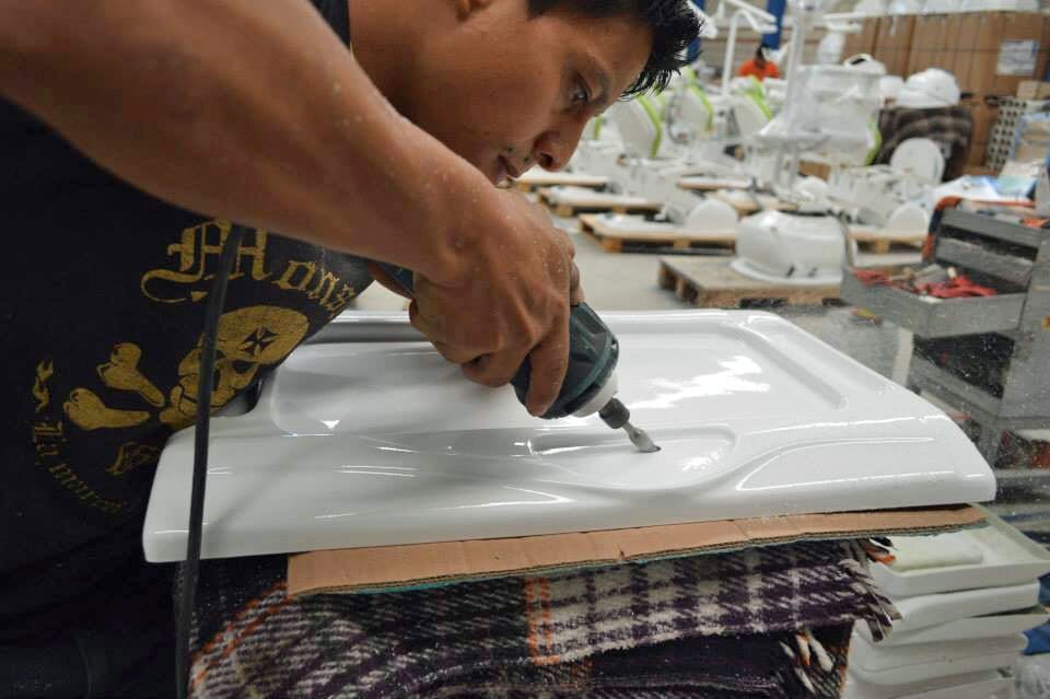 A worker using a drill on a white plastic component in a manufacturing facility, with other parts visible.