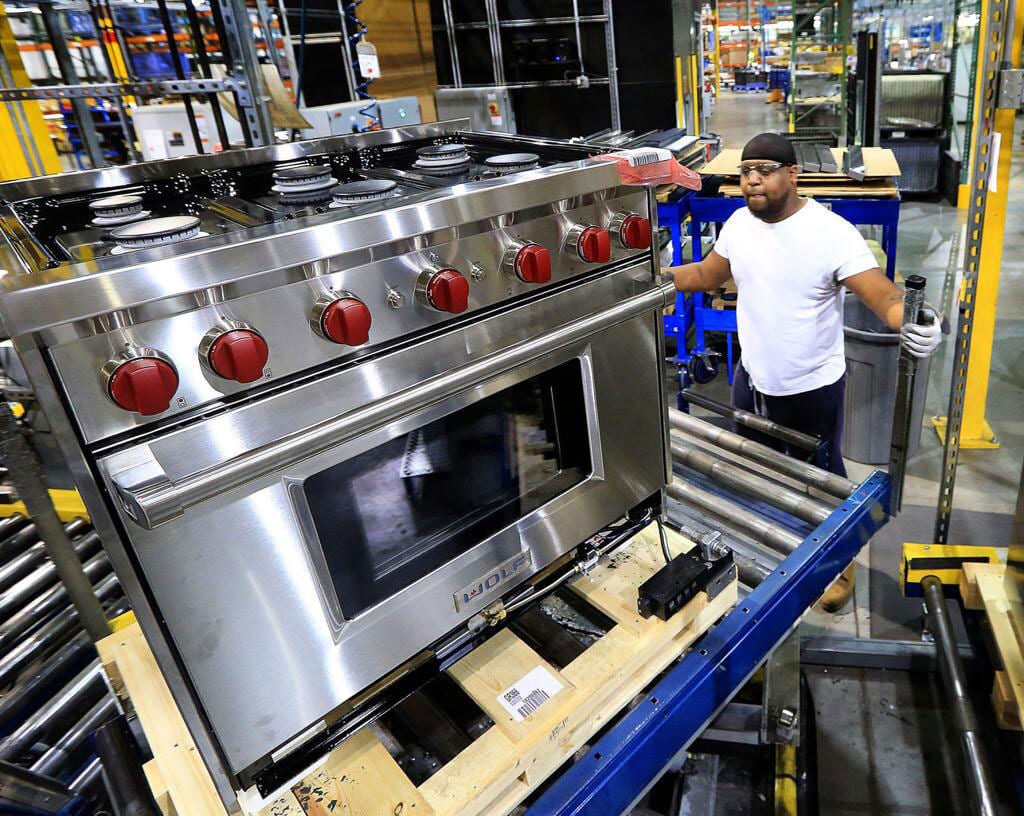Man in a Wolf Appliance factory setting inspecting a shiny stainless steel Wolf range and oven on a conveyor.