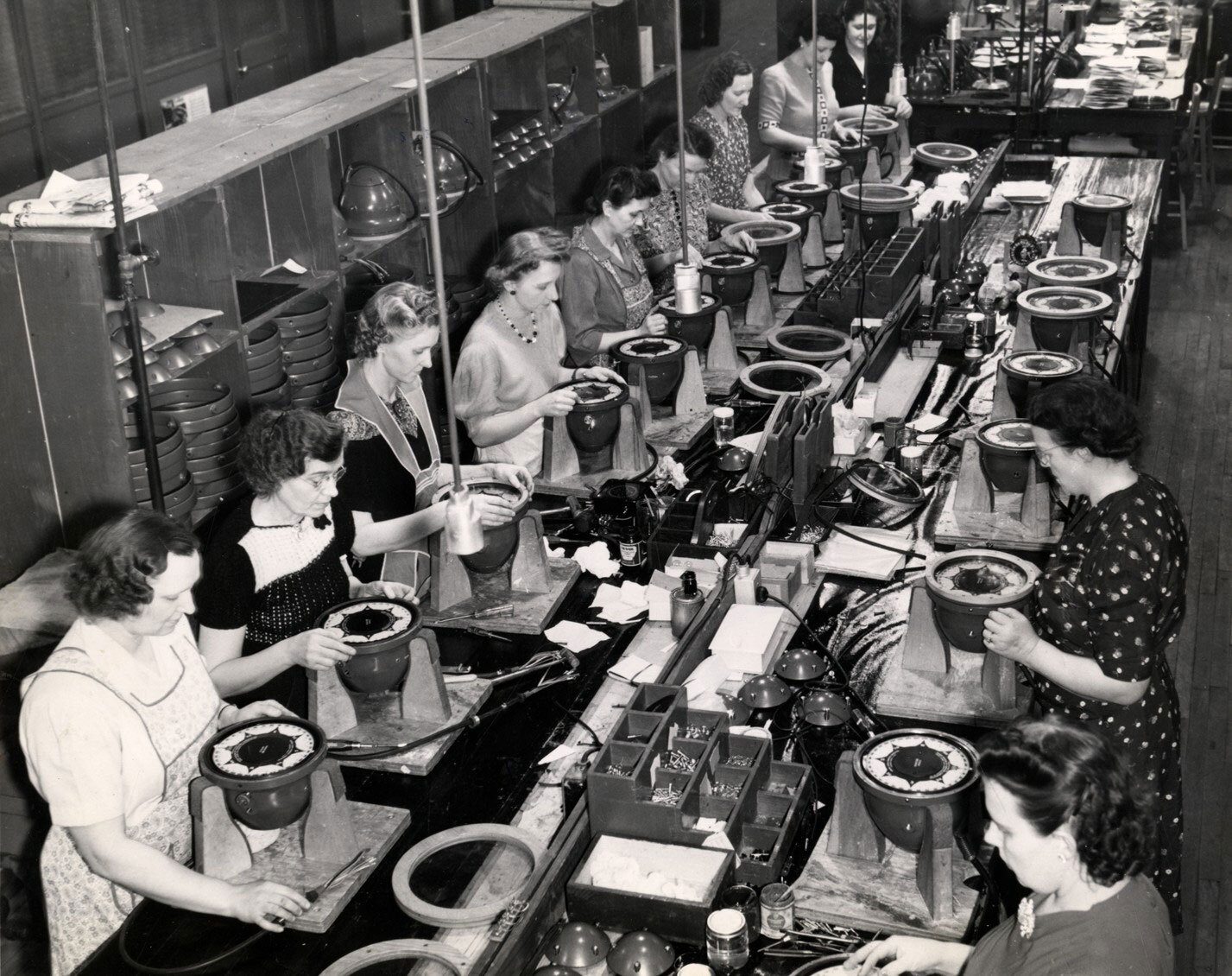 BW photo of women on a factory assembly line, assembling round electrical components.