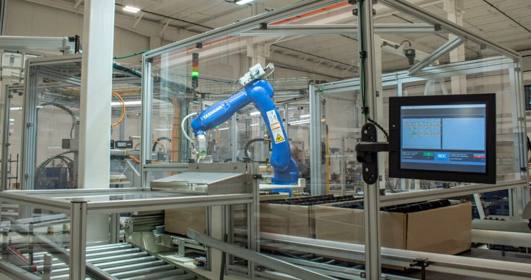 Blue Yaskawa robot arm on an automated production line with conveyor belts, boxes, and a control screen.