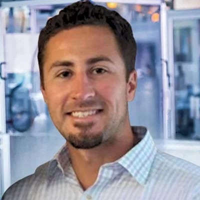 Smiling man with dark hair and beard, wearing a light-colored plaid shirt.