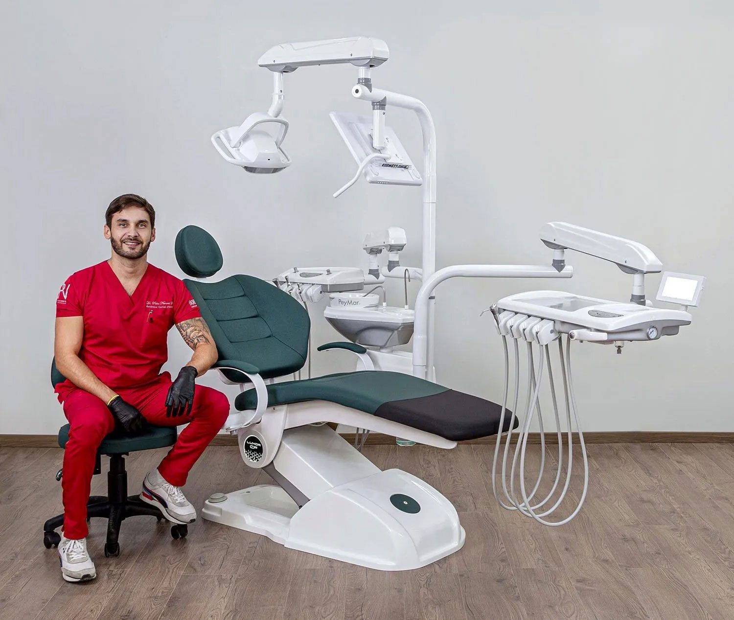 Dentist in red scrubs sitting next to a modern dental chair and equipment in a clean clinic.