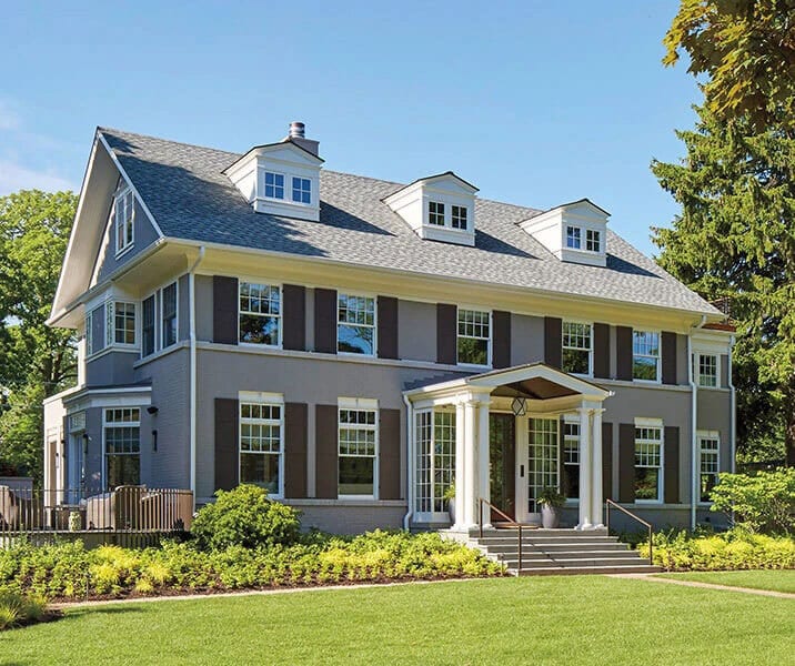 Grey house with dark shutters, white columned porch, and green lawn.