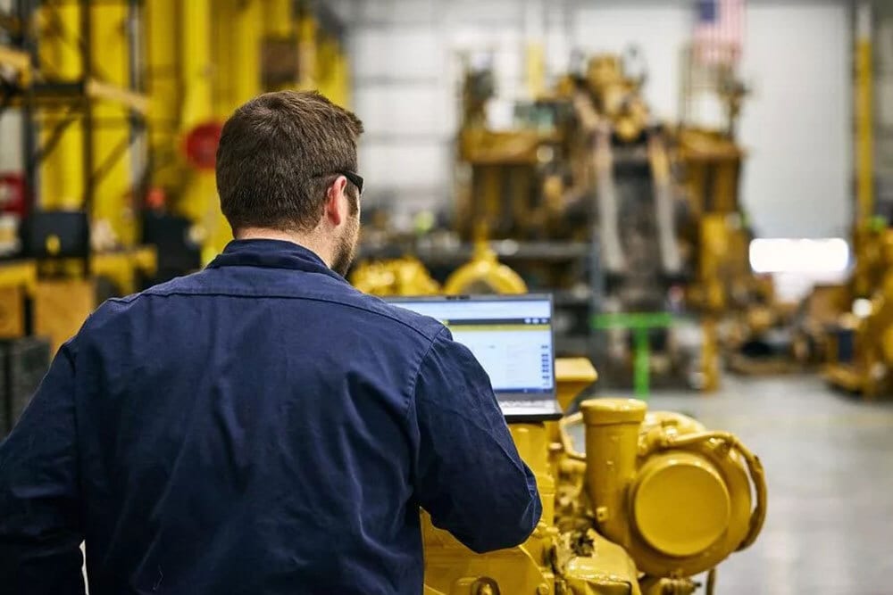 Man in blue shirt with laptop by yellow machinery in an industrial setting.