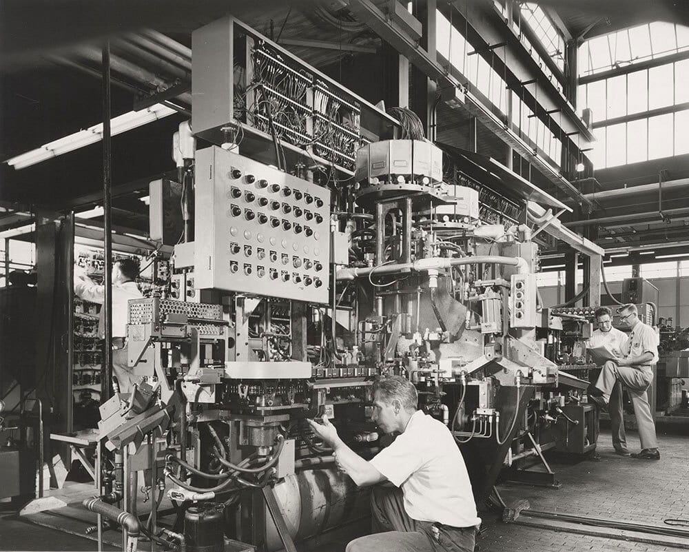 Black and white photo of workers operating and inspecting intricate machinery in a busy factory.
