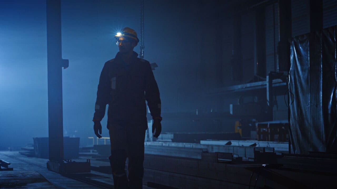 Worker in hard hat with a headlamp walks through a dark, hazy industrial facility.