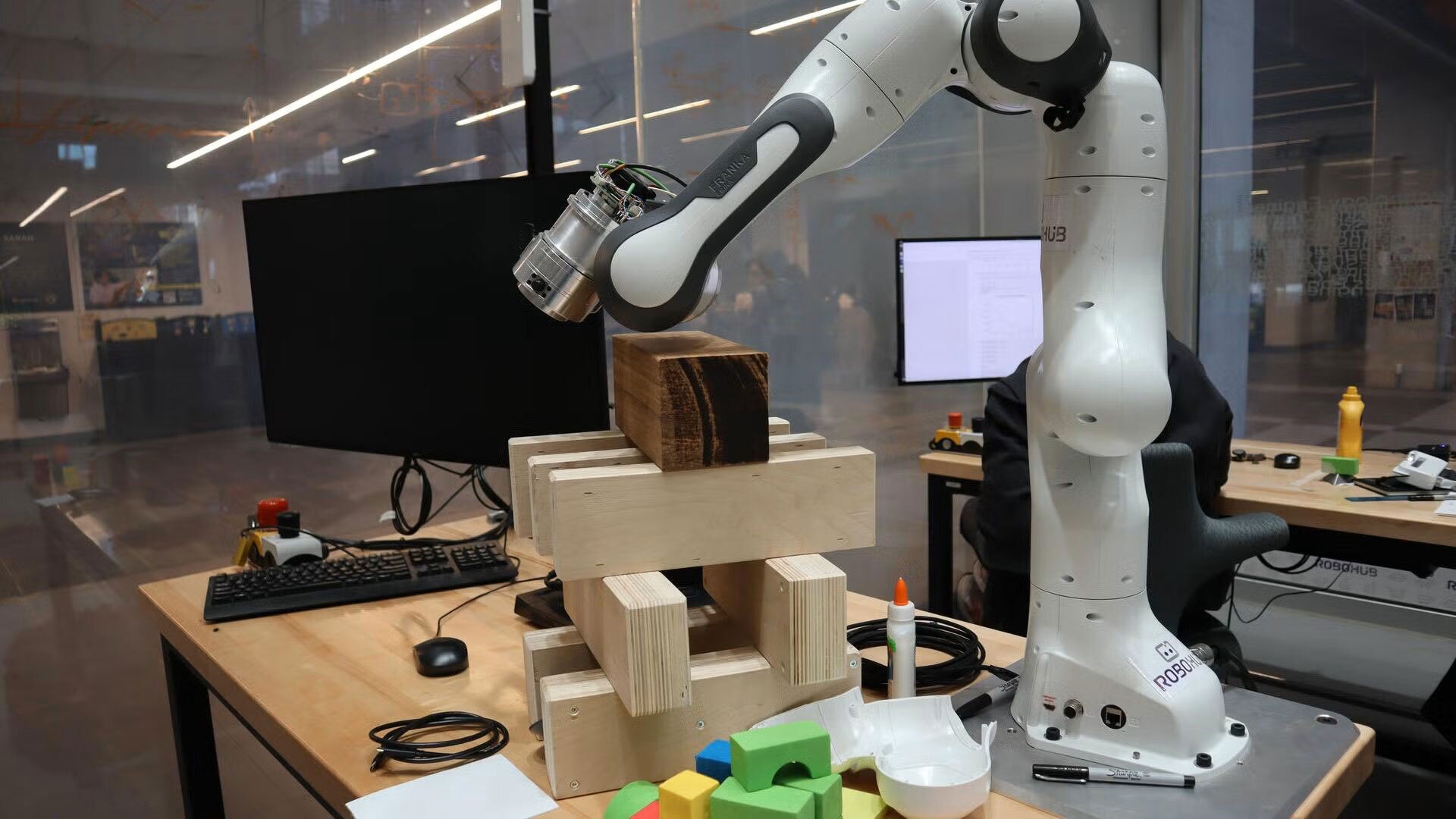 Robotic arm holds a wooden block over a wooden structure on a desk in a lab.
