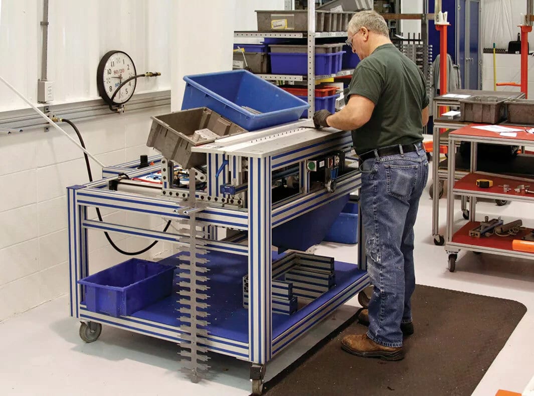 Man assembling metal parts at an industrial workstation with blue bins in a factory.
