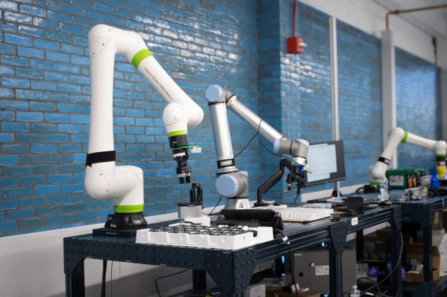 Two robotic arms, one white and one silver, perform tasks on a workbench with microplates against a blue brick wall.