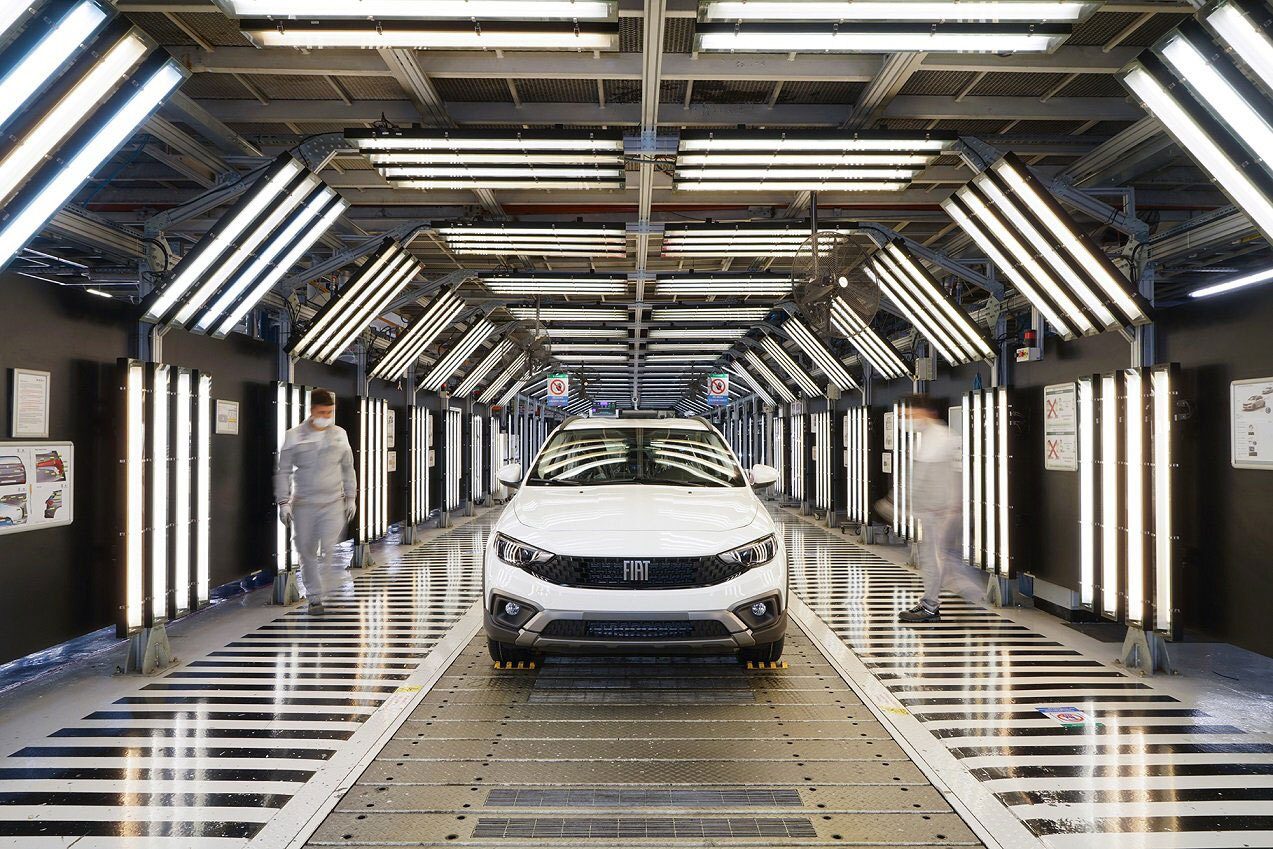 A white Fiat car on a manufacturing inspection line under bright lights with two blurry workers.