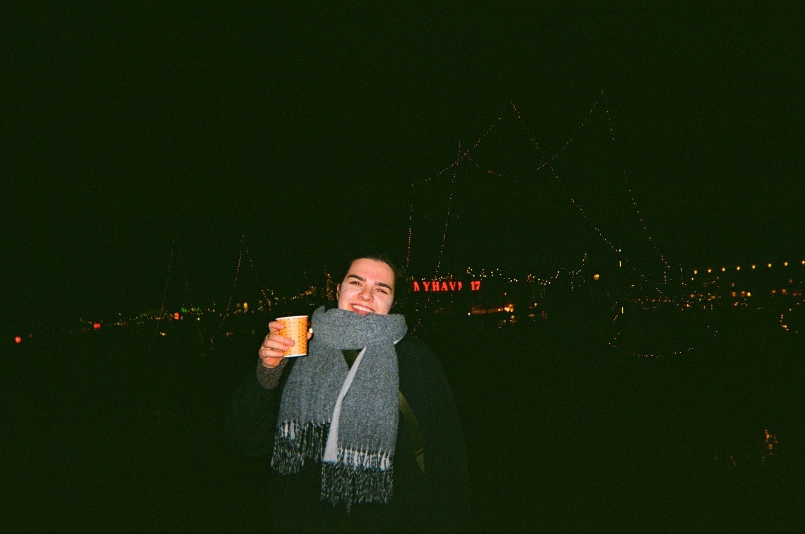 Smiling person in scarf holds a cup at night with blurry Nyhavn 17 background lights.
