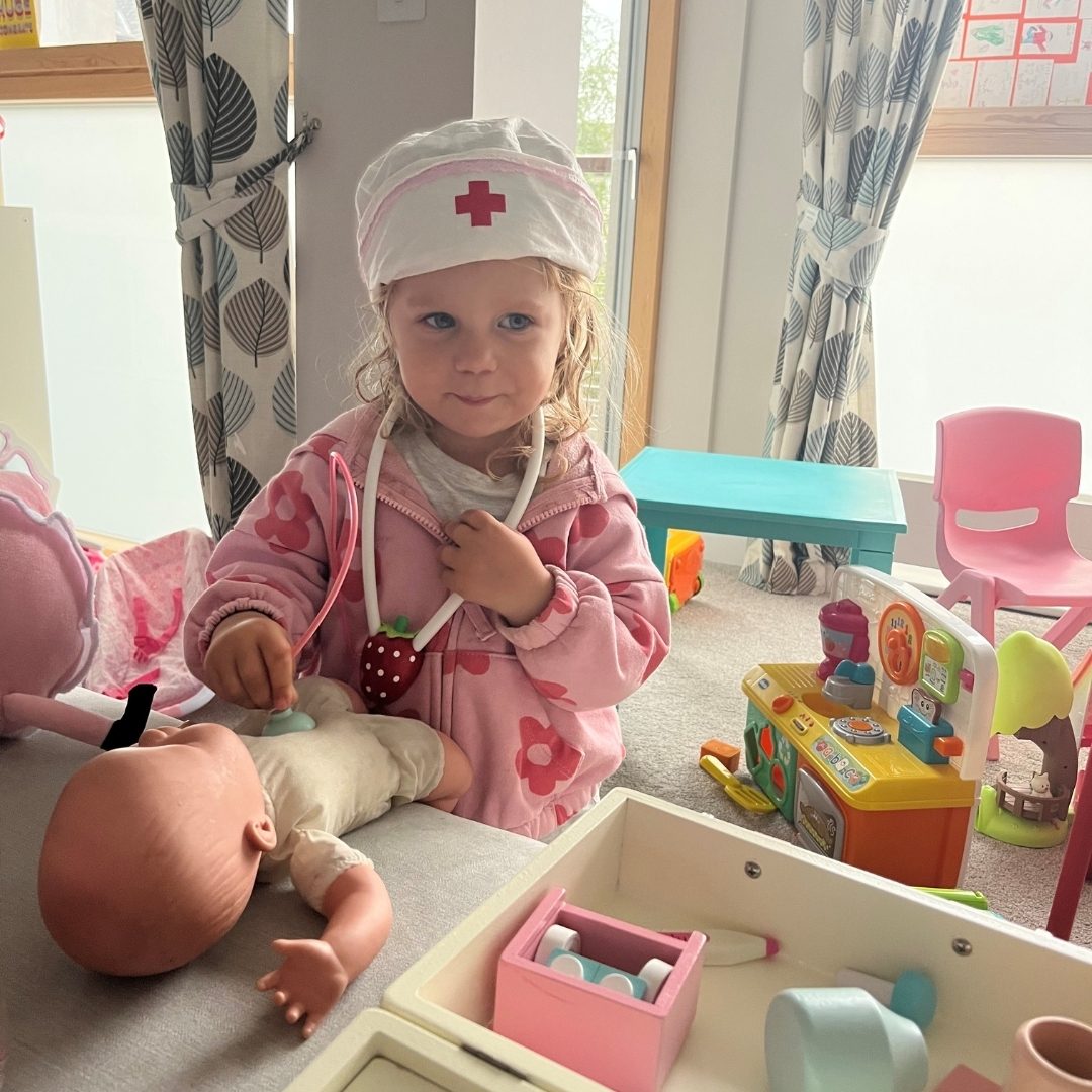 Toddler girl in nurse hat and stethoscope plays doctor with a baby doll.
