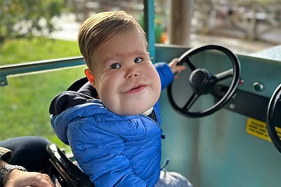 Smiling baby with a round face in a blue jacket, pretending to drive a vehicle.