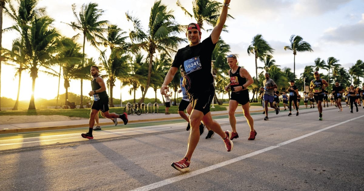 Runners on a palm-lined road during golden hour, one man raises his arm triumphantly.