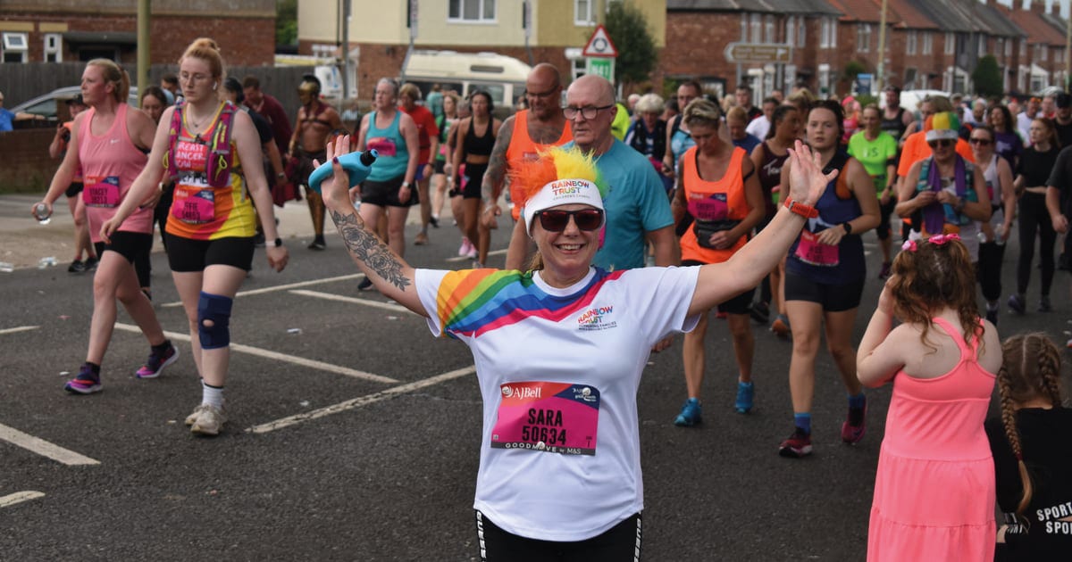 Smiling woman in rainbow shirt and sunglasses, arms outstretched, leading a group in a race.