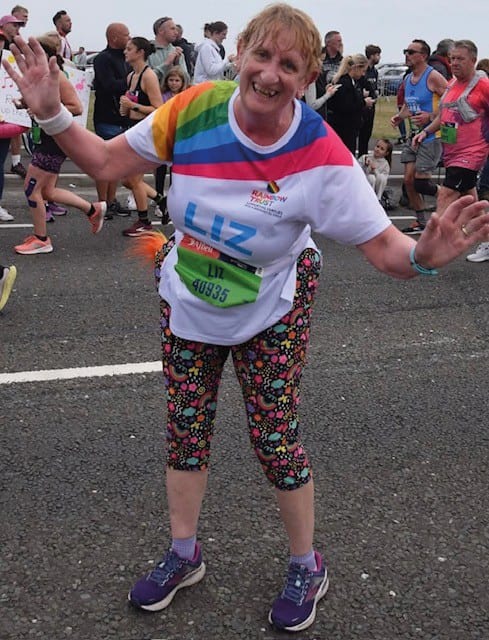 A smiling woman in a rainbow-striped shirt and colorful patterned leggings waves during a race.