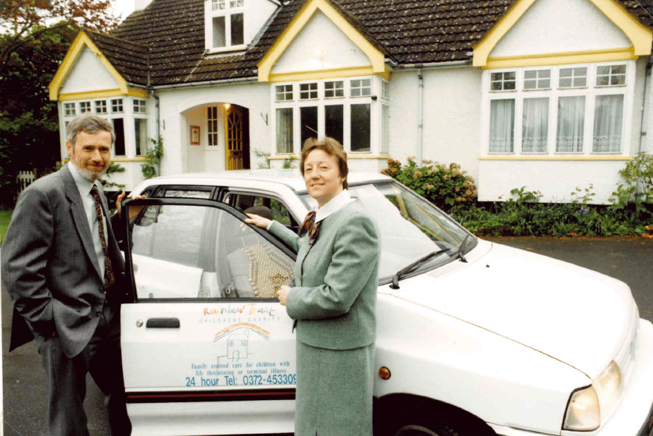 Two people, a man and a woman, stand by a white Rainbow Trust charity car in front of a house.