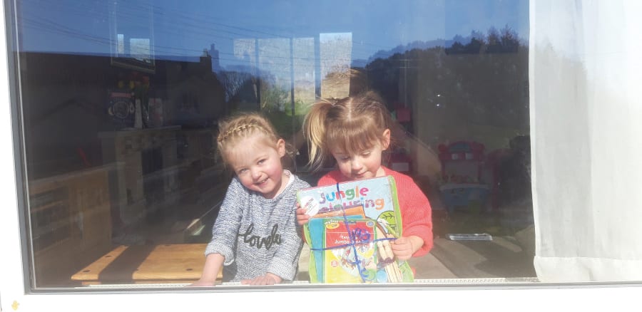 Two smiling young girls seen through a window, one holding a stack of books and colouring supplies.
