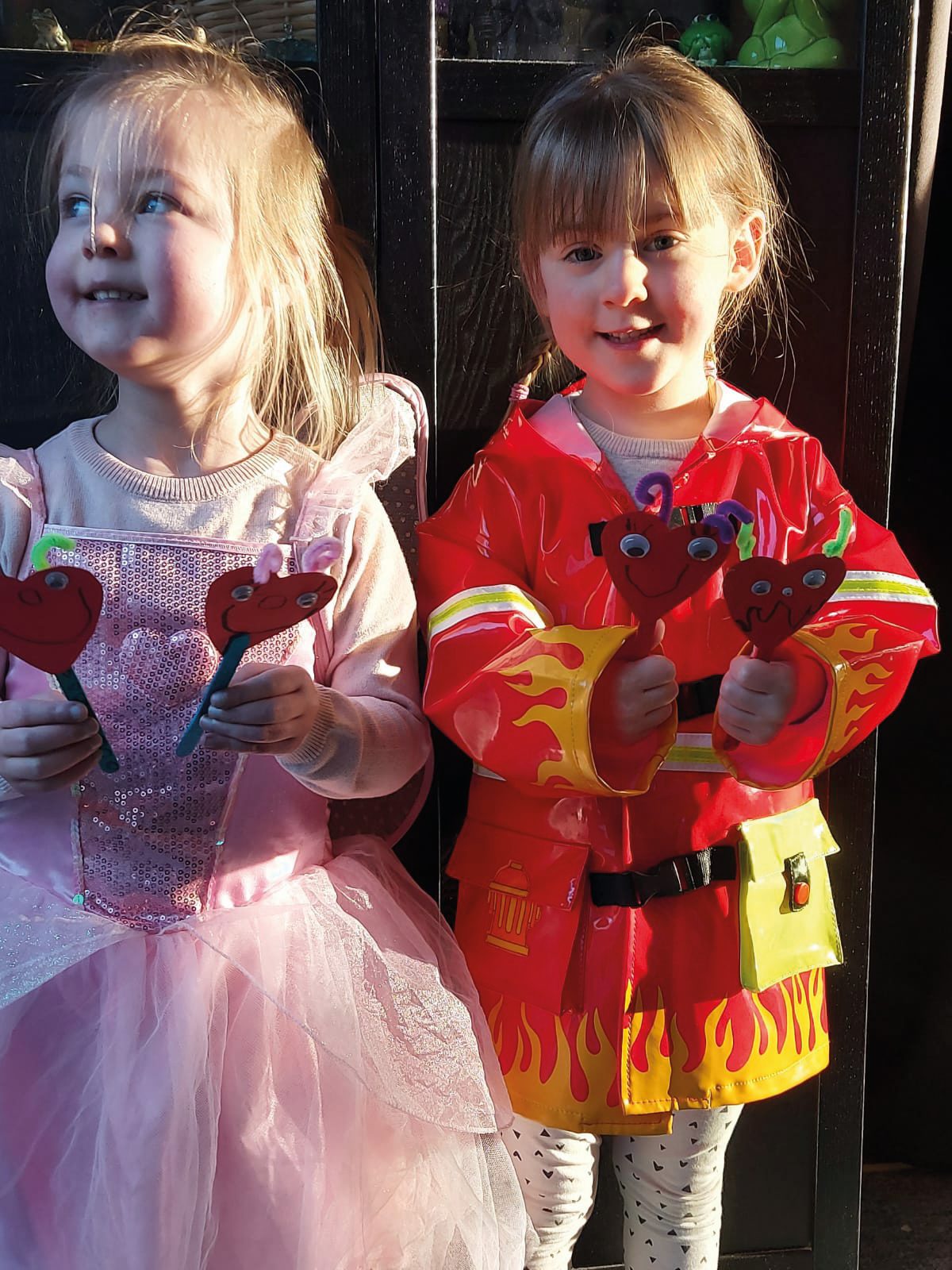 Two young girls in costumes, one a princess and one a firefighter, holding heart-shaped crafts.