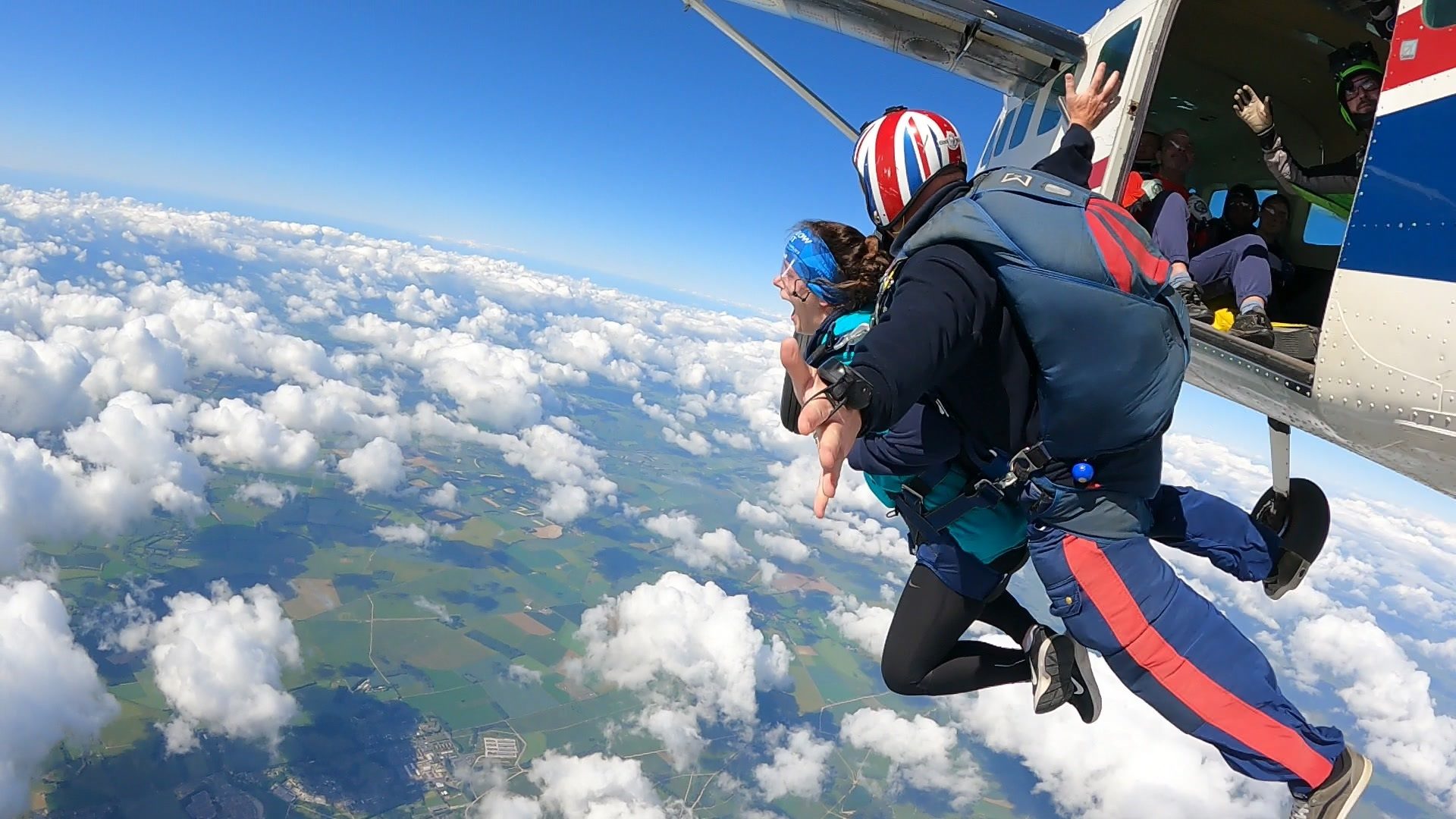 Two skydivers exit an airplane high above clouds and green landscape, one with an excited expression.