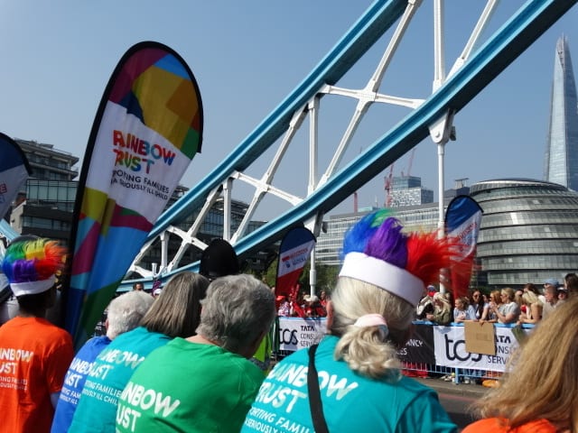 People in "Rainbow Trust" shirts and rainbow wigs at an event, with Tower Bridge, The Shard, and City Hall.