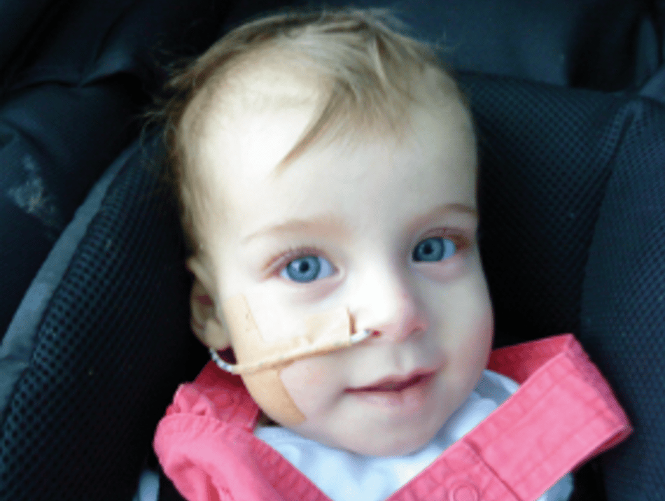 Close-up of a baby with blue eyes, light hair, and a feeding tube, smiling in a car seat.