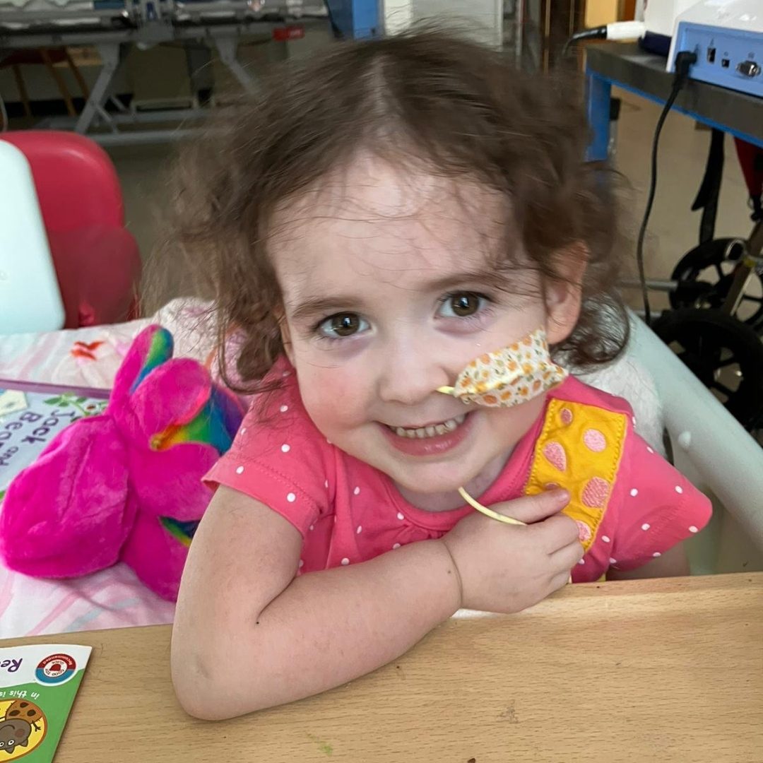 Smiling young girl with an NG tube, wearing a pink shirt, looks at the camera.