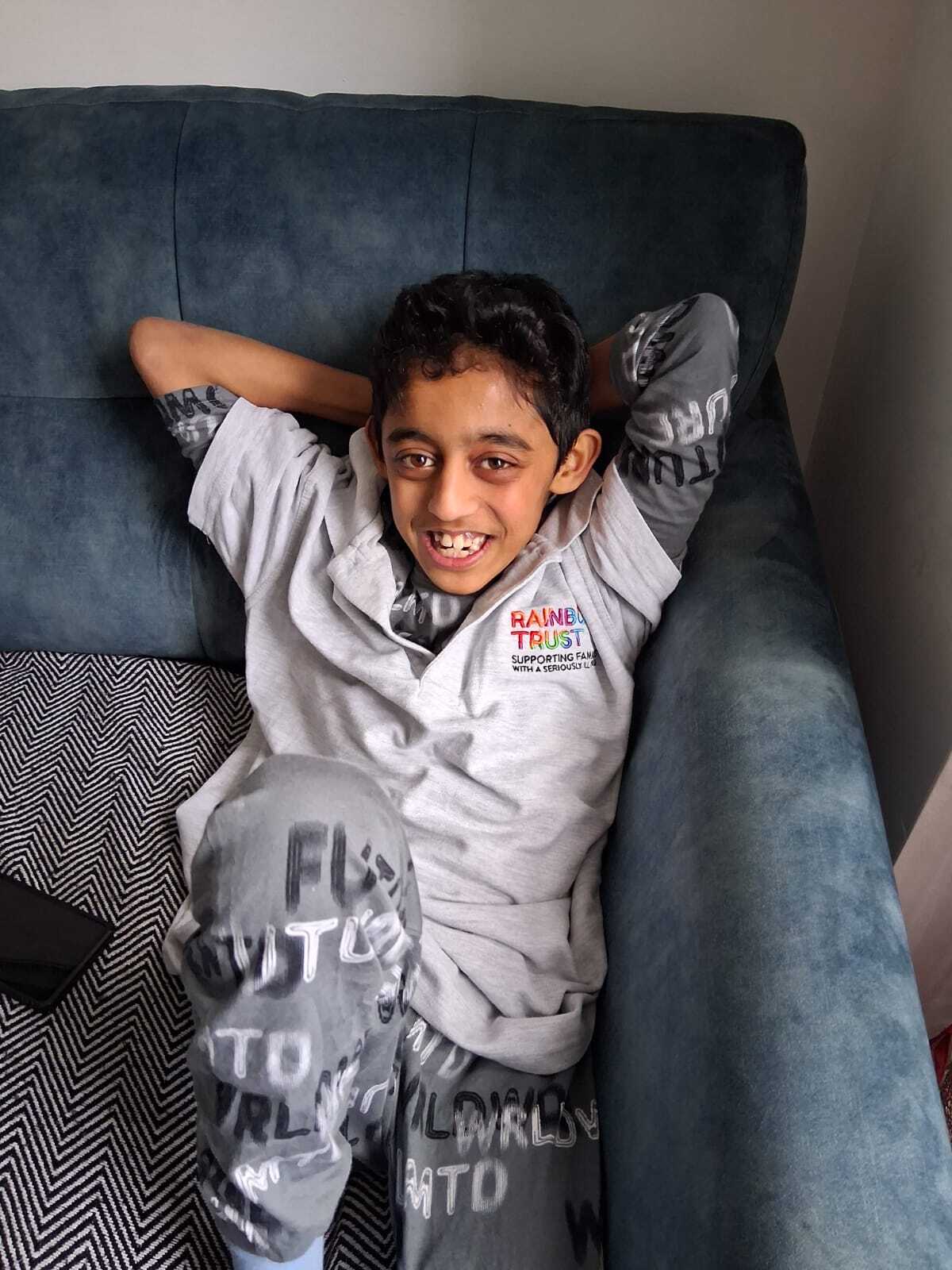 Smiling boy in gray 'Rainbow Trust' hoodie and pants, lying on a blue sofa, arms behind head.