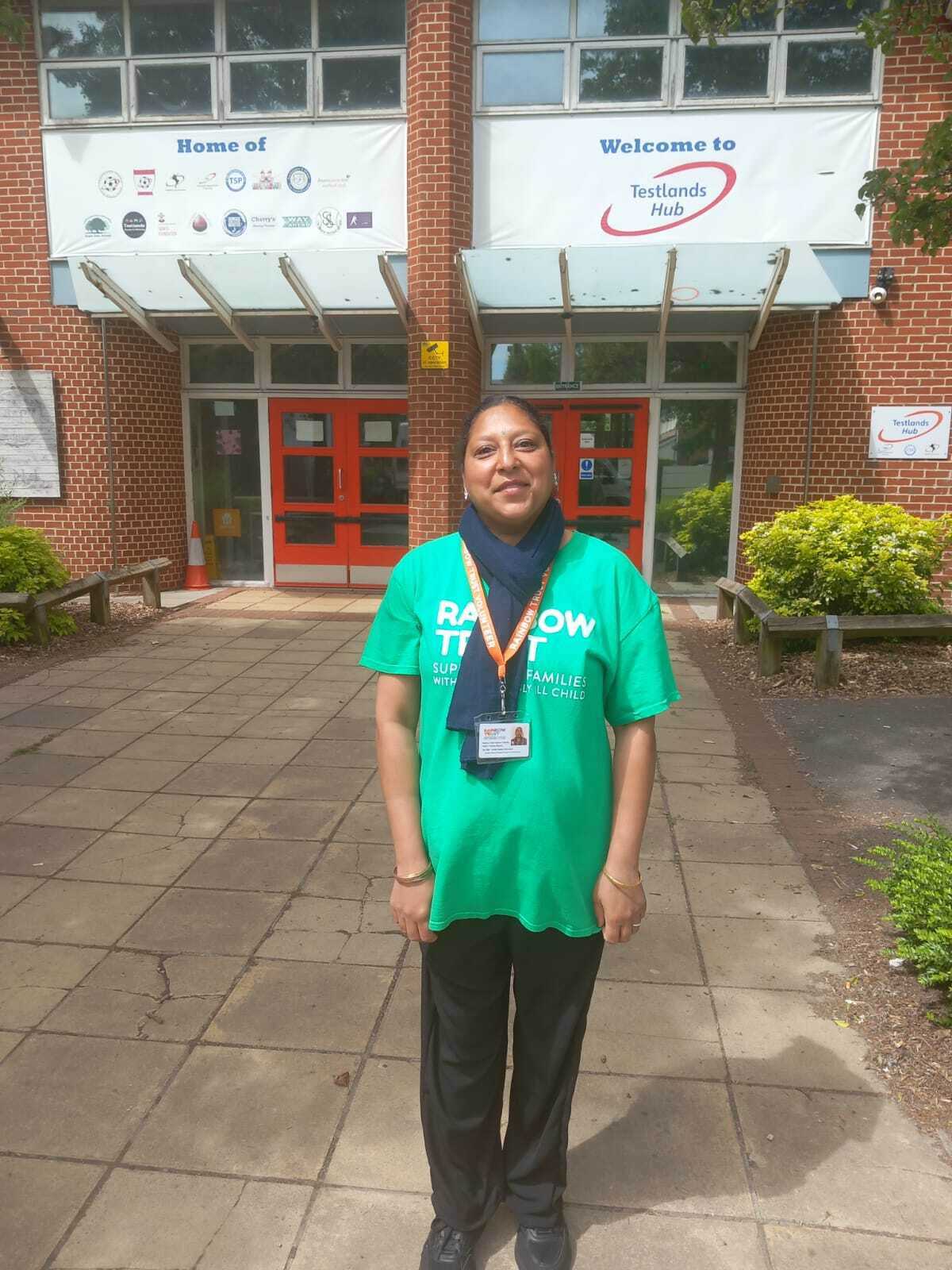 Woman in green 'Rainbow Trust' t-shirt stands outside a brick building with 'Testlands Hub' banner.