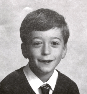 Black and white portrait of a young boy with short hair, wearing a uniform and tie, smiling faintly.