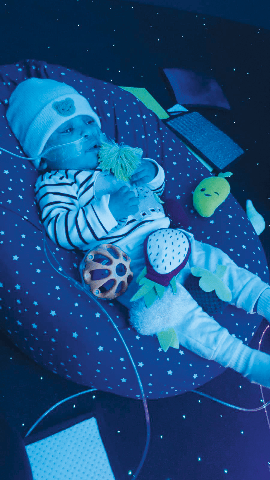 Baby with nasal tube, toys on starry cushion in dark blue room with glowing stars.
