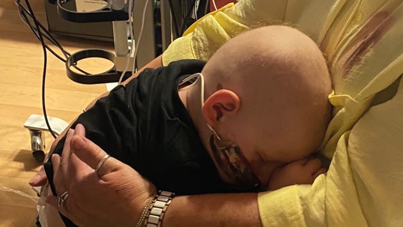 A bald child with medical tubes is embraced by an adult in a hospital setting.