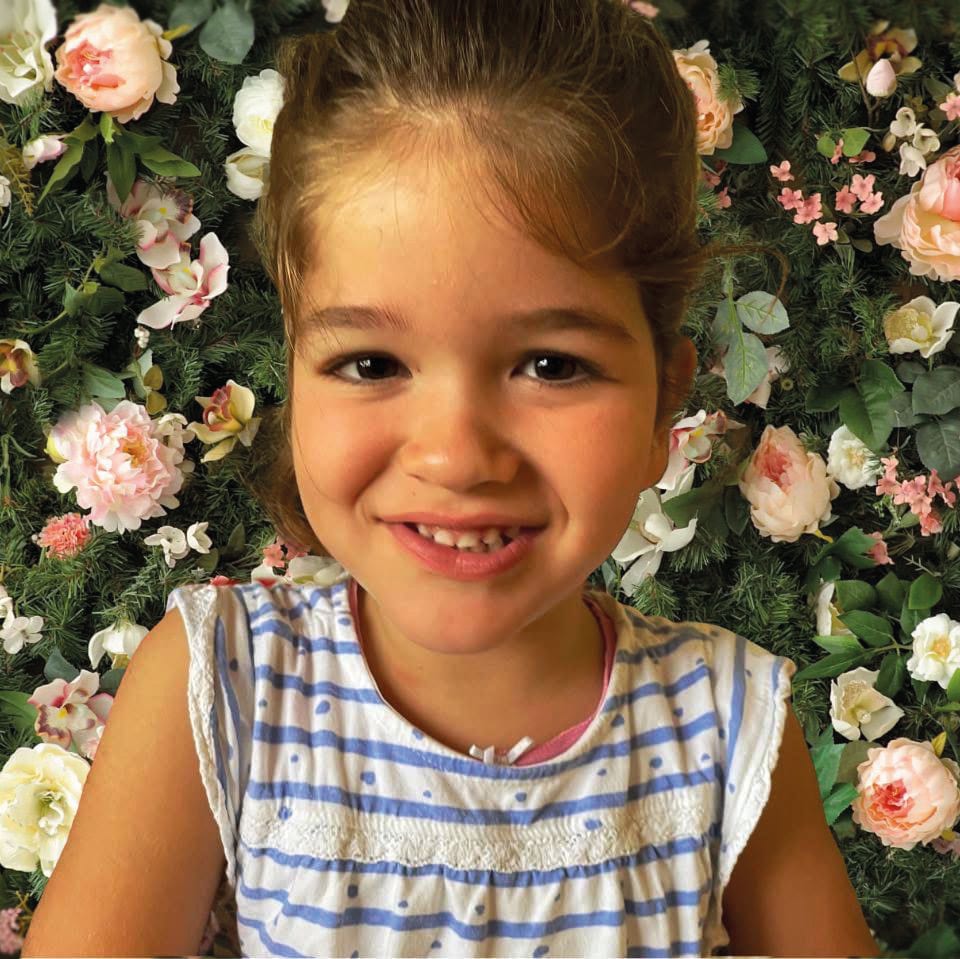 Smiling young girl in a striped top against a wall of pink and white flowers and green foliage.