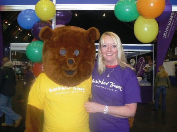 Bear mascot and woman in purple "Rainbow Trust" shirt pose smiling amongst colorful balloons indoors.