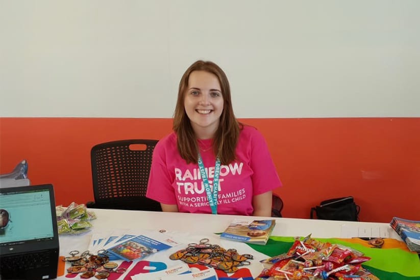 Smiling woman in pink "Rainbow Trust" shirt behind a table with merchandise.